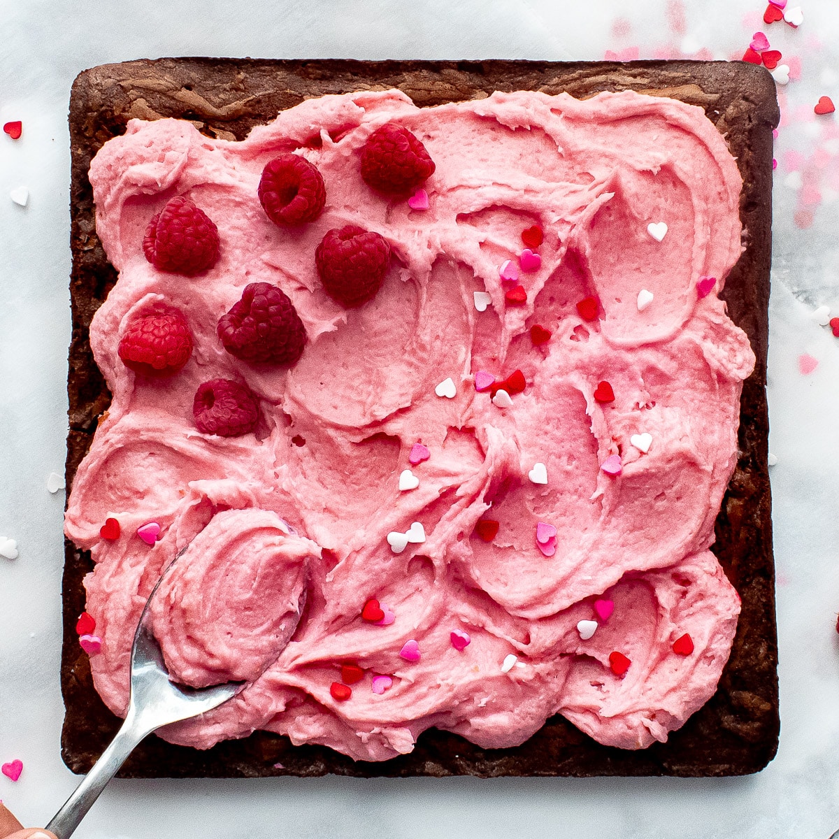 A spoon spreading frosting on raspberry chocolate brownies with fresh raspberries and sprinkles