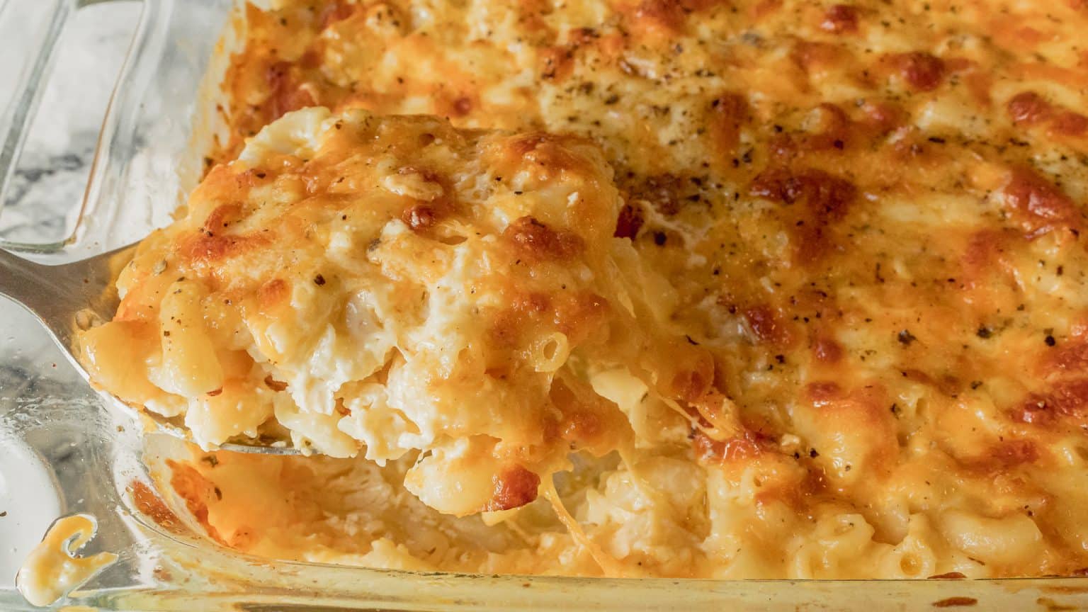 A close-up of cheesy baked macaroni and cheese in a glass dish. A spatula is lifting a serving, showing creamy, gooey, melted cheese coating the pasta. The top is browned and crispy, sprinkled with seasoning.