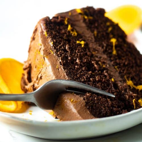slice of dark chocolate orange cake being cut with a fork, with an orange and orange zest as garnish