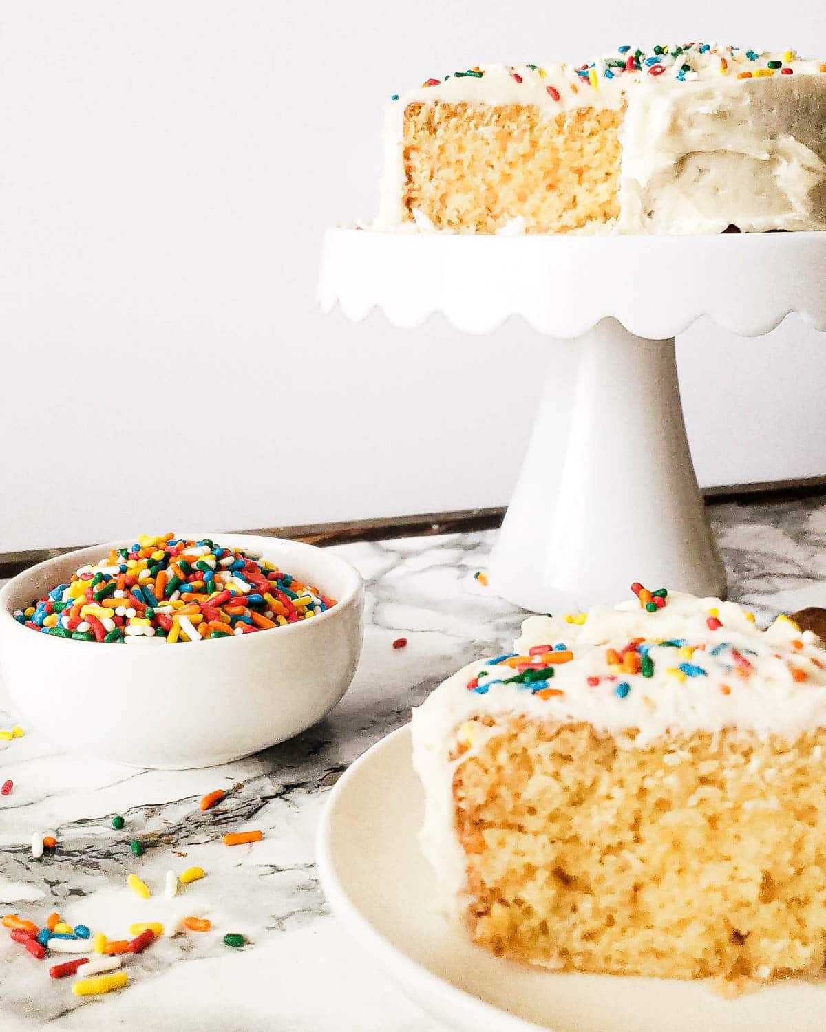 mini vanilla cake on a cake stand with a bowl of sprinkles and a slice of cake