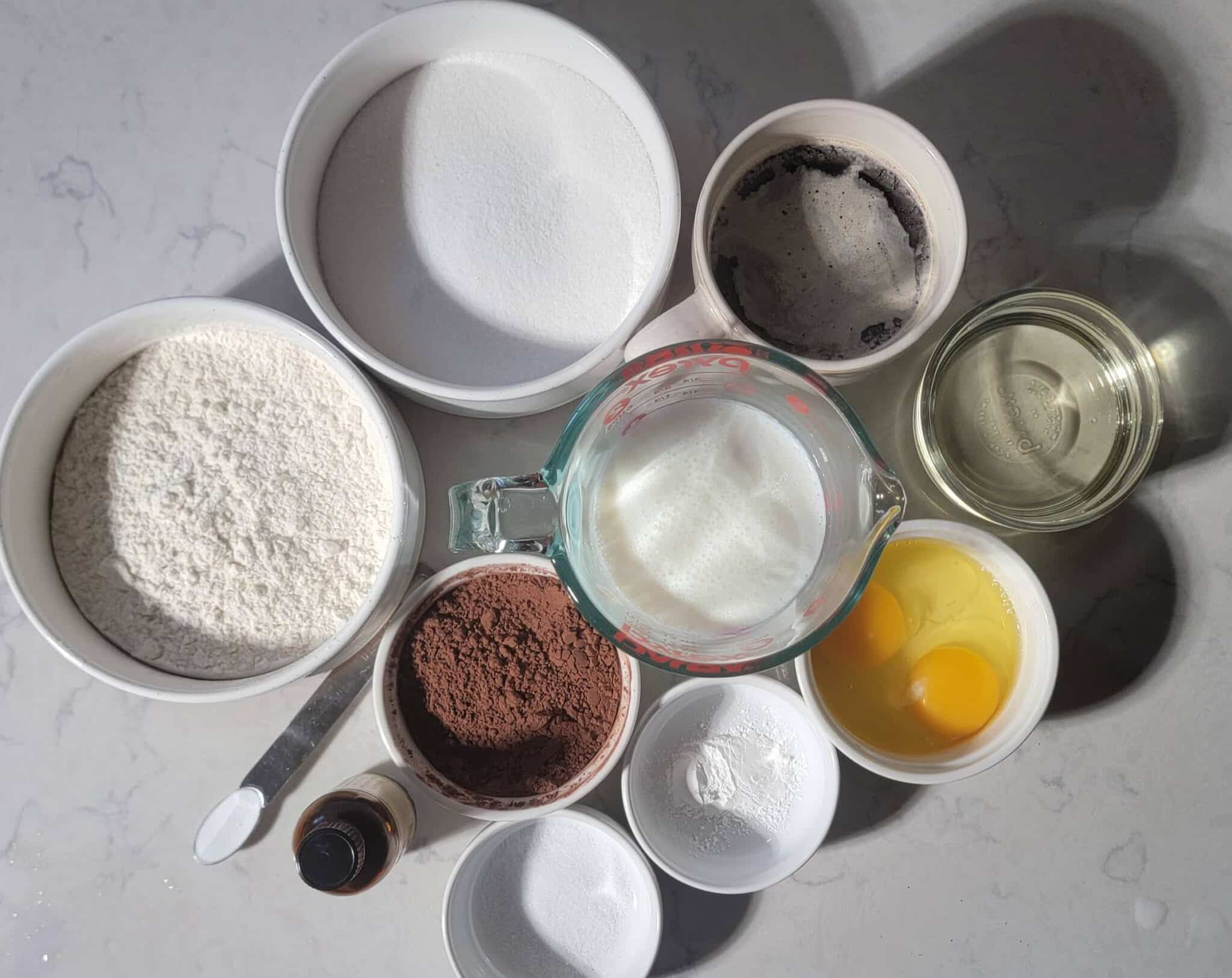 Ingredients for Matilda's Chocolate Cake recipe neatly laid out on a marble countertop, including flour, sugar, cocoa powder, and eggs, with sunlight casting soft shadows.