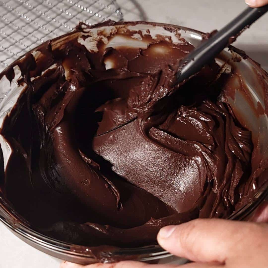 Stirring a shiny, fudge frosting in a glass bowl for creating the perfect Matilda's Chocolate Cake.