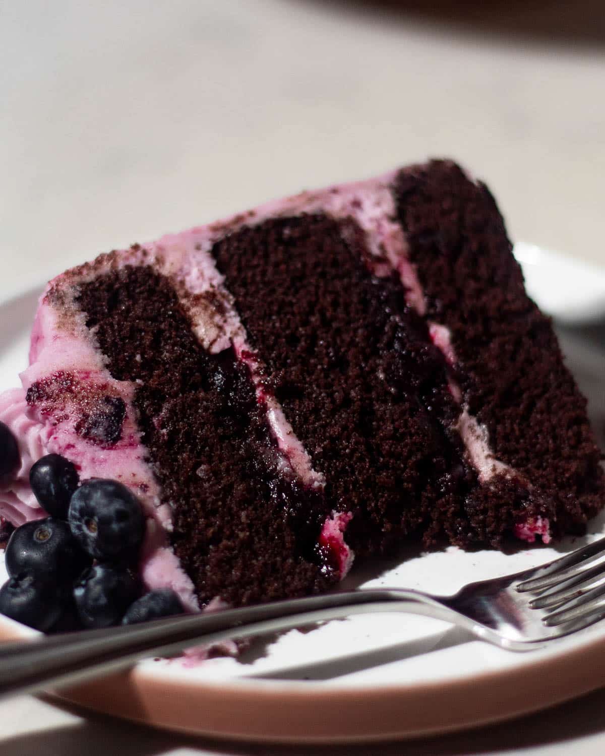 Tall slice of chocolate blueberry cake layered with dark chocolate sponge, blueberry filling, and pink berry frosting.