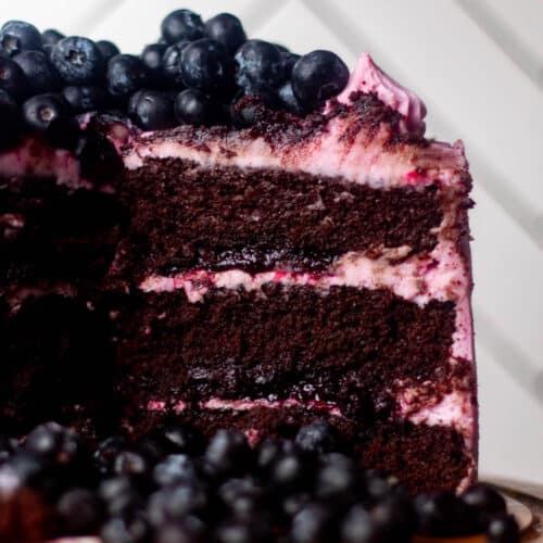 Close-up of a chocolate blueberry cake slice showing rich chocolate layers, thick blueberry filling, and swirled berry frosting topped with fresh blueberries.