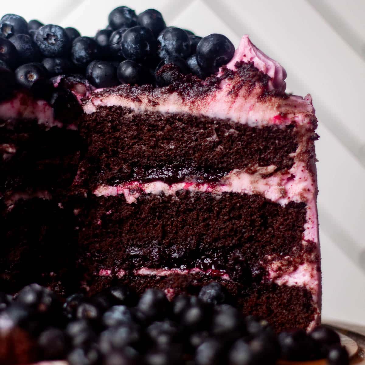 Close-up of a chocolate blueberry cake slice showing rich chocolate layers, thick blueberry filling, and swirled berry frosting topped with fresh blueberries.
