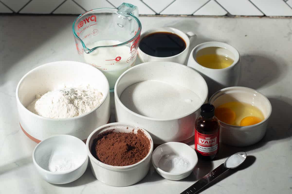 Ingredients for chocolate blueberry cake on a countertop—flour, sugar, cocoa powder, baking soda, eggs, milk, oil, coffee, and vanilla extract.
