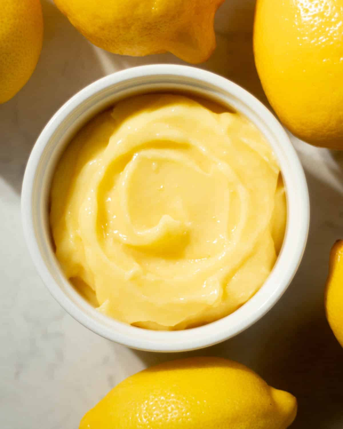 Top-down view of silky lemon curd in a white dish, encircled by fresh yellow lemons—perfect for a simple lemon curd recipe.