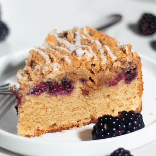 A single slice of blackberry coffee cake served on a white plate with fresh blackberries. The golden cake is topped with streusel and a drizzle of vanilla glaze.