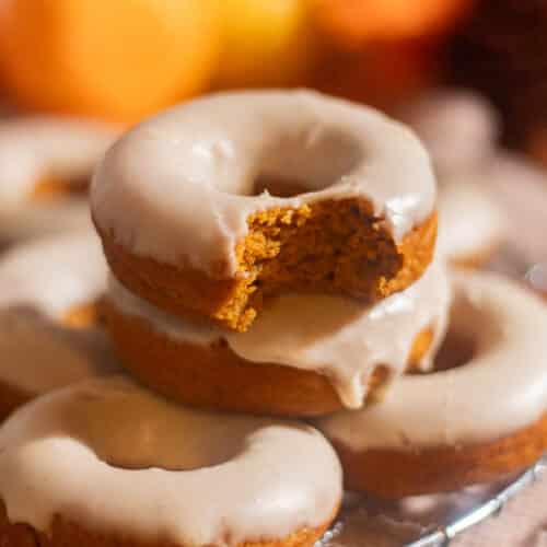 Square image of a bitten pumpkin spice donut stacked on others, highlighting its rich, tender crumb and creamy glaze.