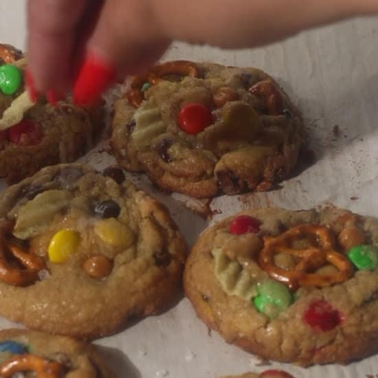 Hand sprinkling sea salt over freshly baked kitchen sink cookies topped with pretzels, M&Ms, potato chips, and chocolate chips.