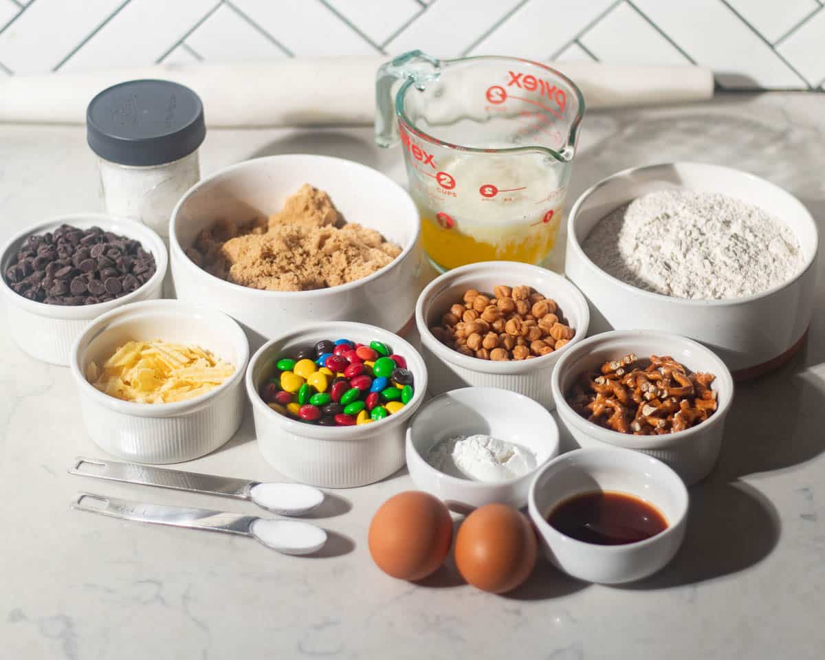 Overhead shot of kitchen sink cookie ingredients including flour, brown sugar, melted butter, chocolate chips, M&Ms, pretzels, potato chips, peanut butter chips, eggs, vanilla, baking soda, and salt arranged on a marble counter.