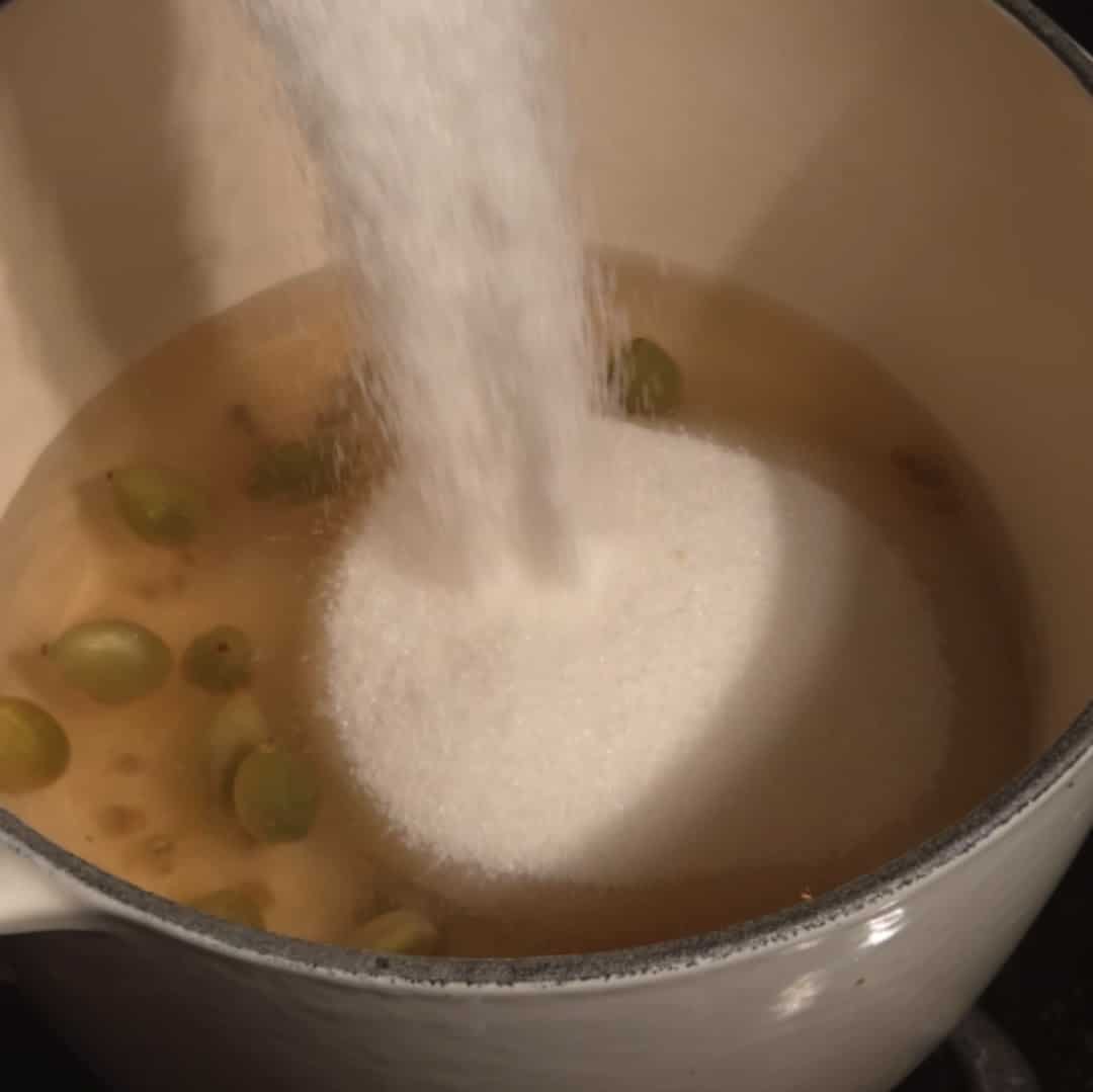 Pouring sugar into simmering water with cardamom pods to dissolve and create cardamom simple syrup.