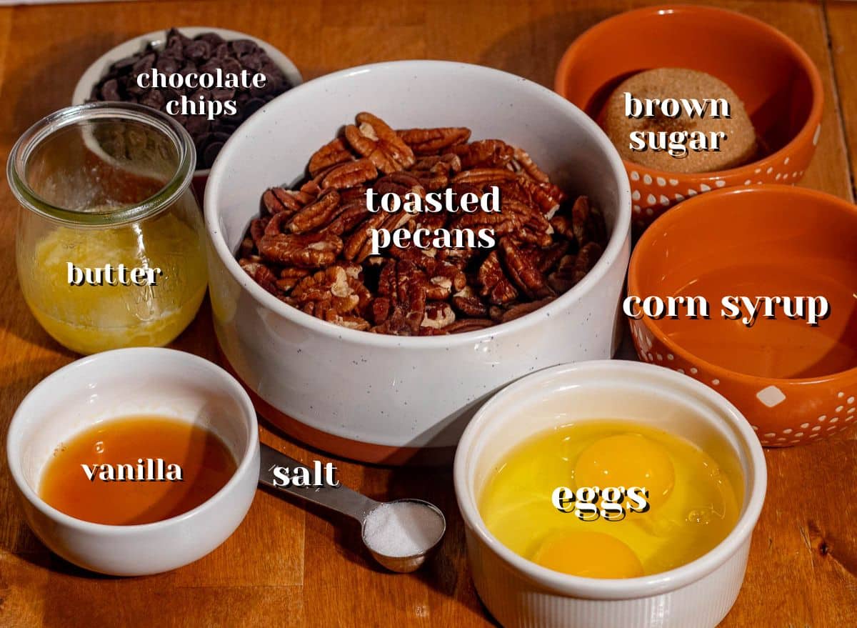 Ingredients for chocolate pecan pie bars including brown sugar, corn syrup, eggs, butter, toasted pecans, and chocolate chips on a wooden table.