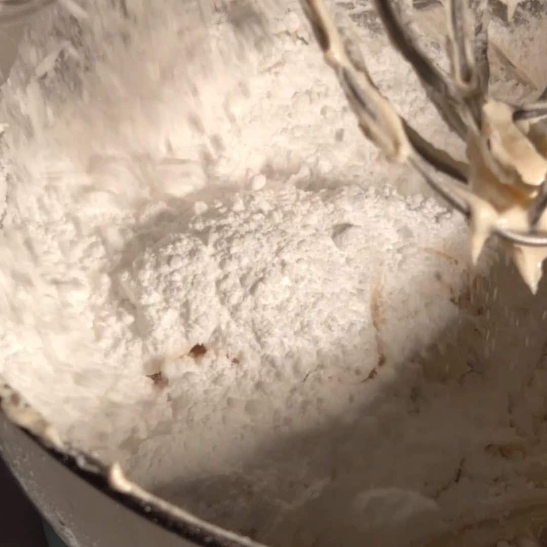 Powdered sugar being added to brown butter cream cheese frosting in a mixing bowl during the frosting mixing process.